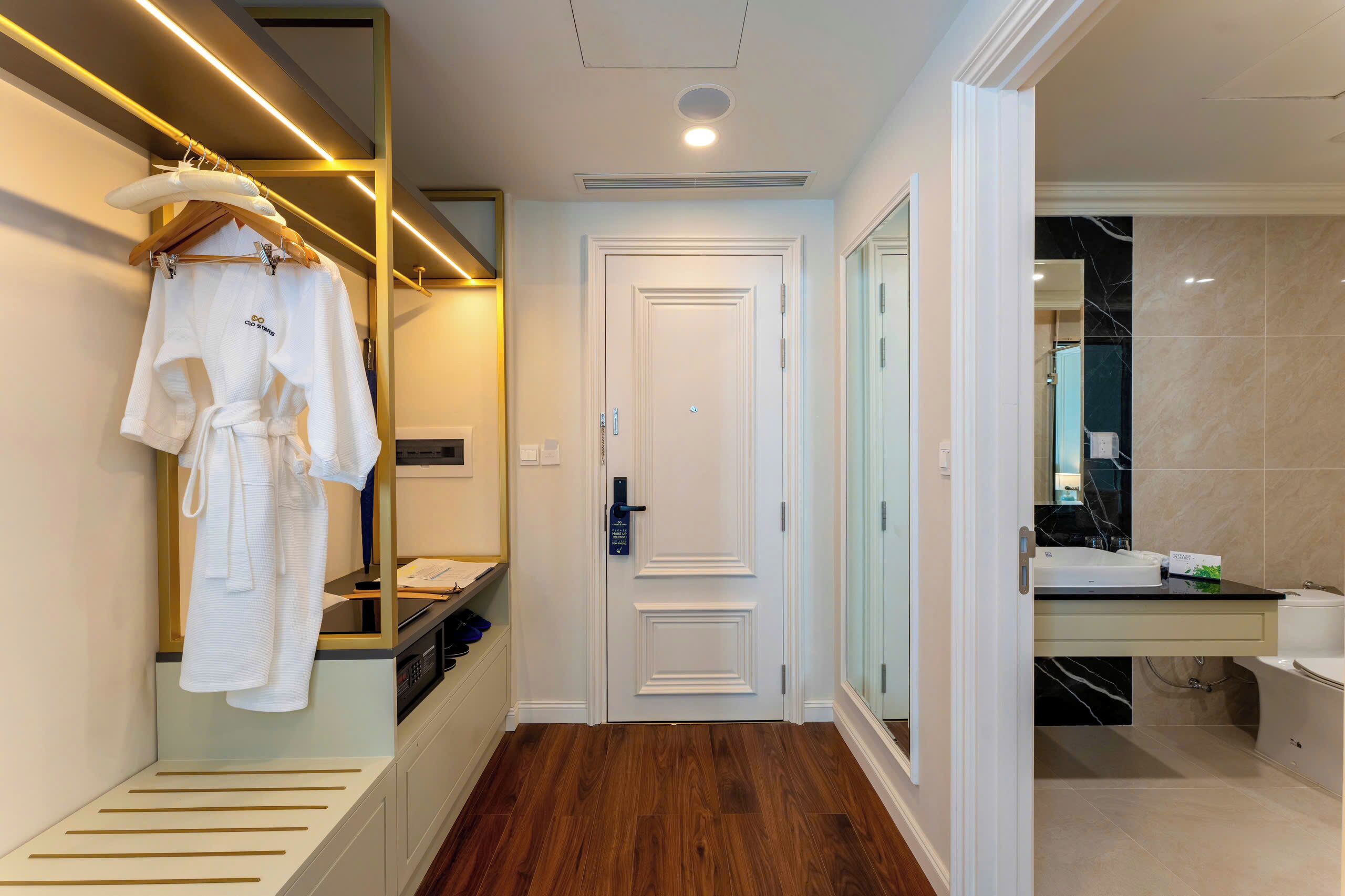 A hotel room with a wooden floor, a white door, a mirror, and a bathroom with a sink and toilet.