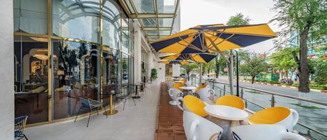 A covered outdoor seating area with tables and chairs, a glass storefront, and a view of a river.