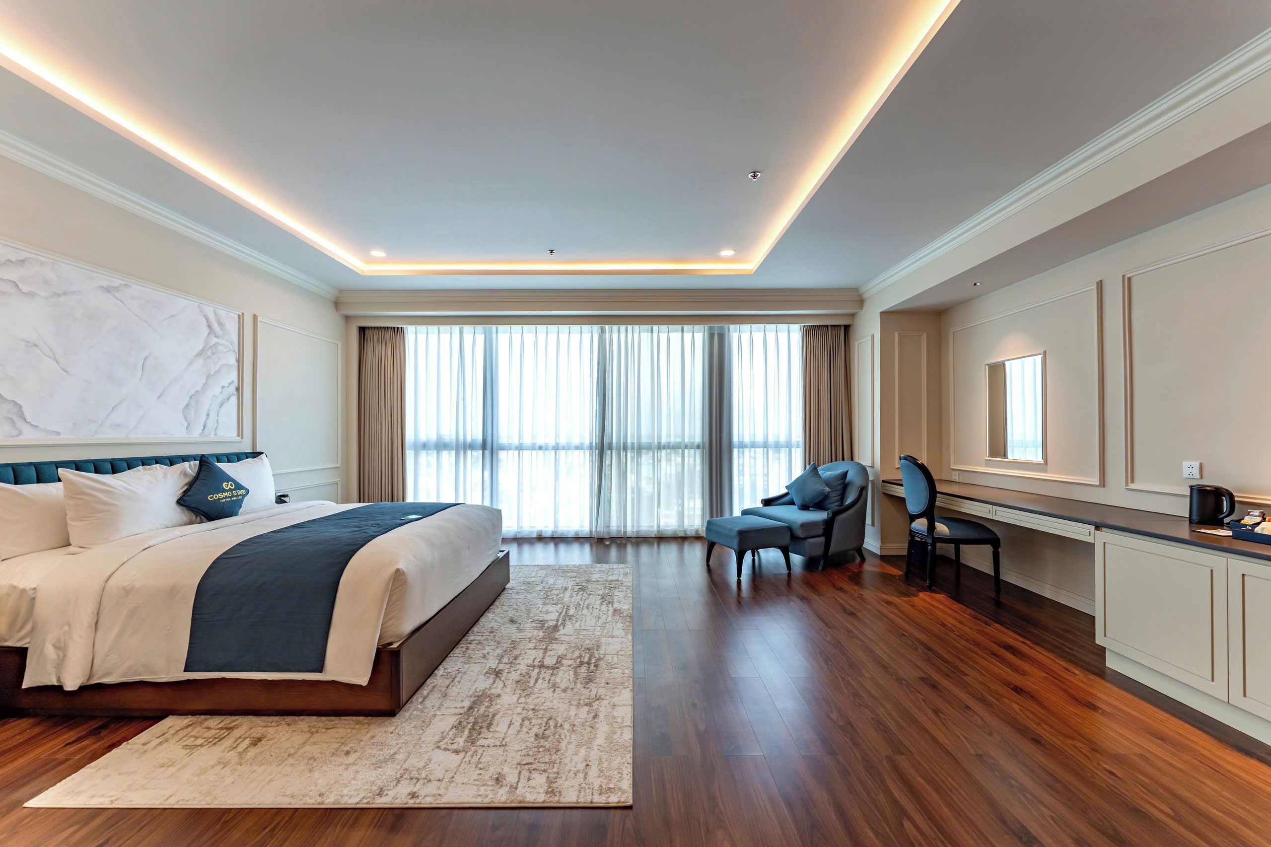 A modern hotel room with a large bed, a desk, and a view of the city through large windows.