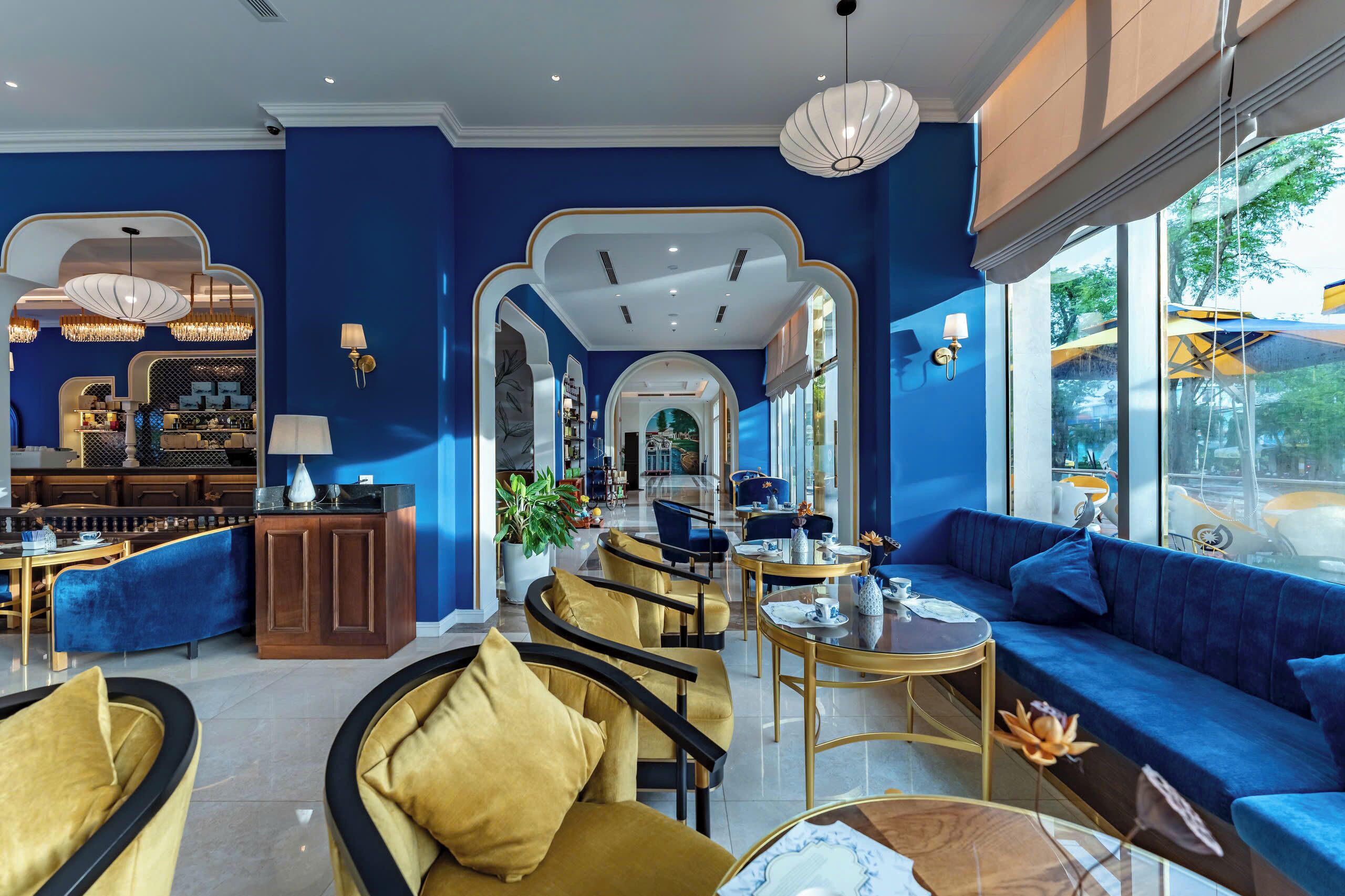 A modern hotel lobby with a blue color scheme, featuring a seating area with sofas and chairs, a bar, and a large mirror reflecting the space.