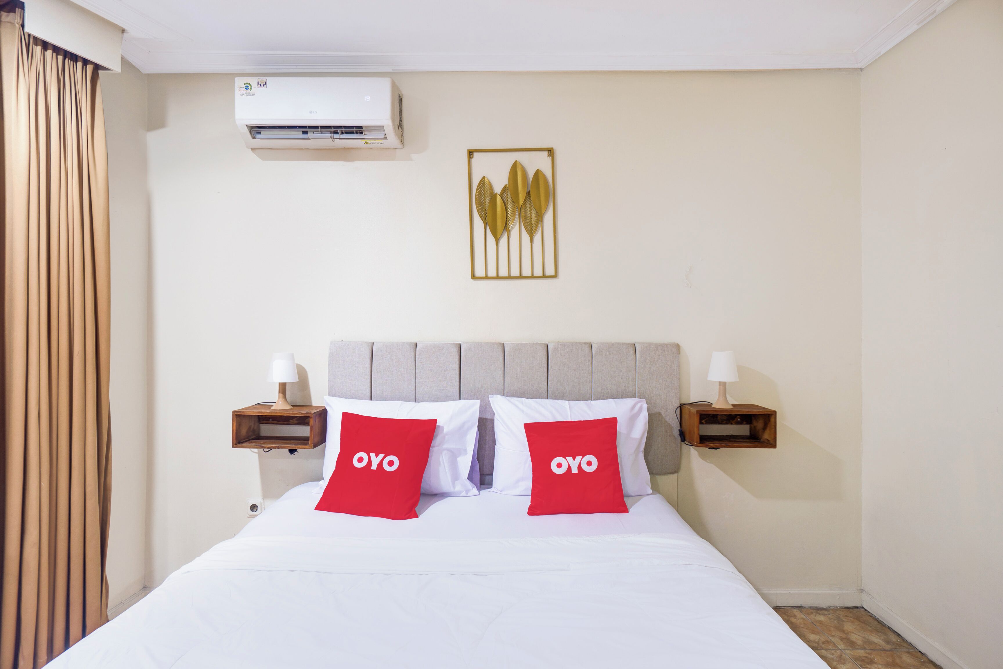 Una habitación de hotel con una cama, dos almohadas rojas con la palabra 'OYO' y un aire acondicionado de pared, además de un adorno en la pared sobre la cama.