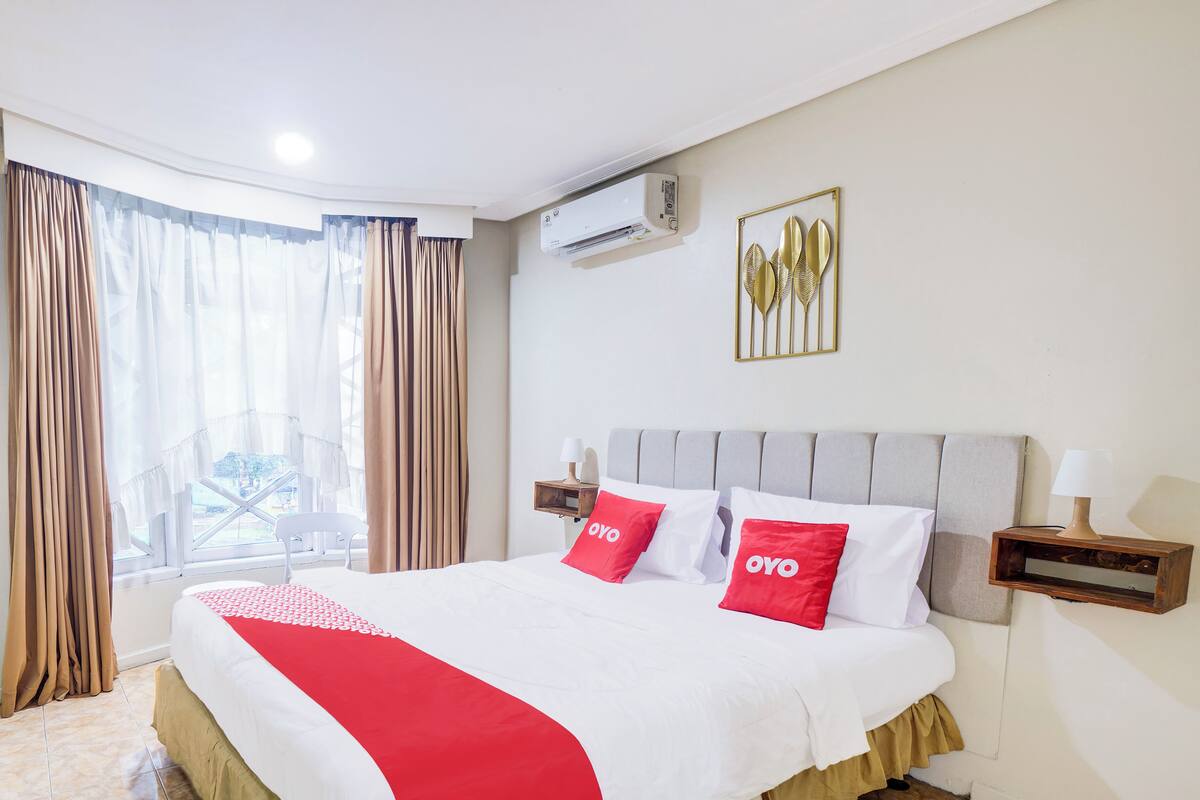 Une chambre d’hôtel avec un lit, des rideaux, un climatiseur, une lampe fixée au mur et une fenêtre donnant sur un paysage agréable.