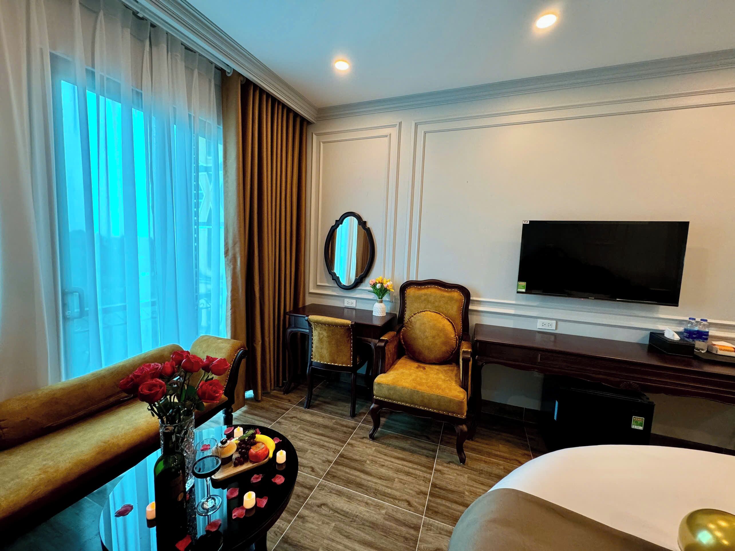 A hotel room with a bed, a desk with a chair, a TV, a window with curtains, and a vase with flowers.