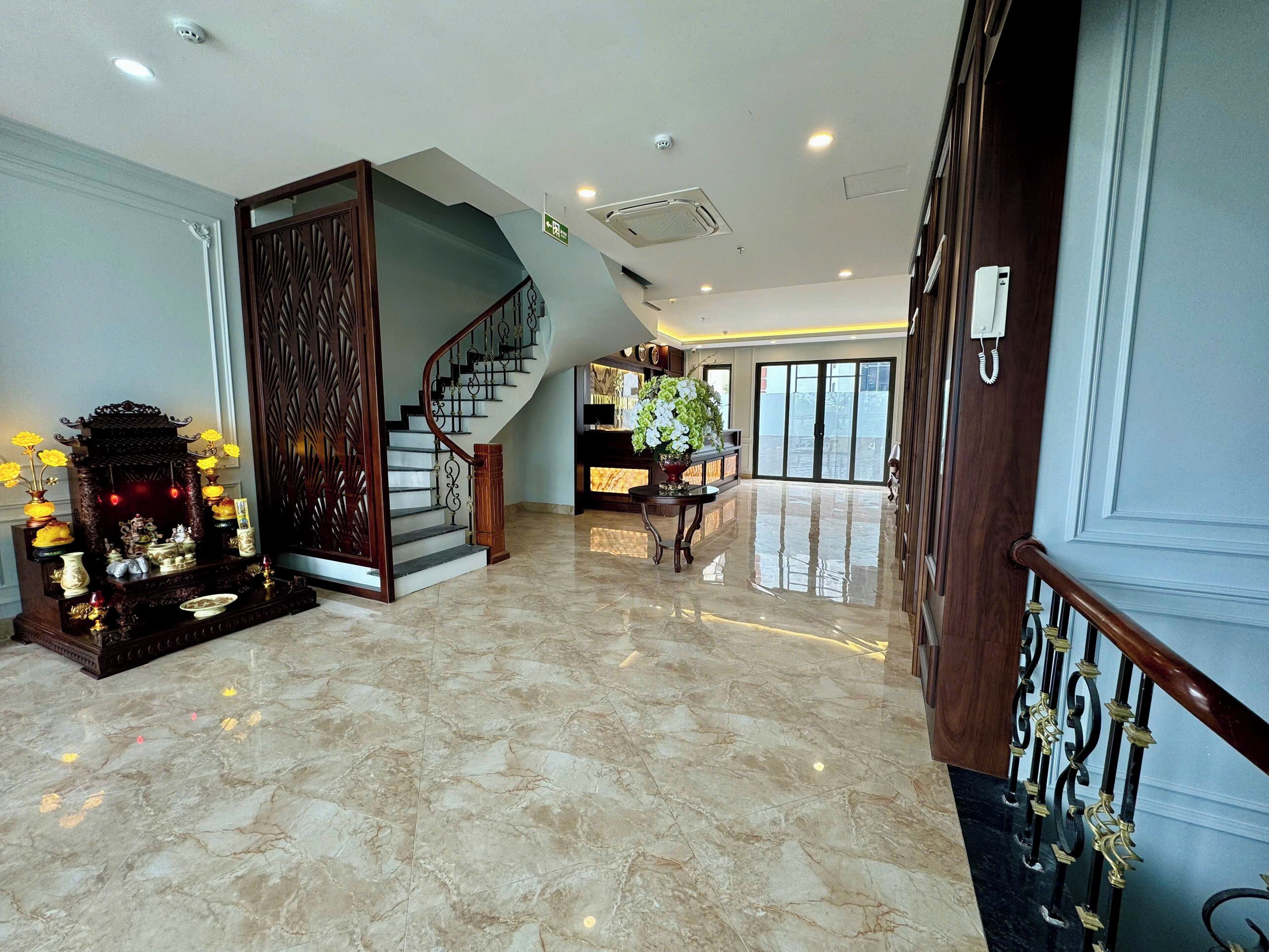 A spacious lobby with a marble floor, a wooden staircase, and a reception desk with a large potted plant.