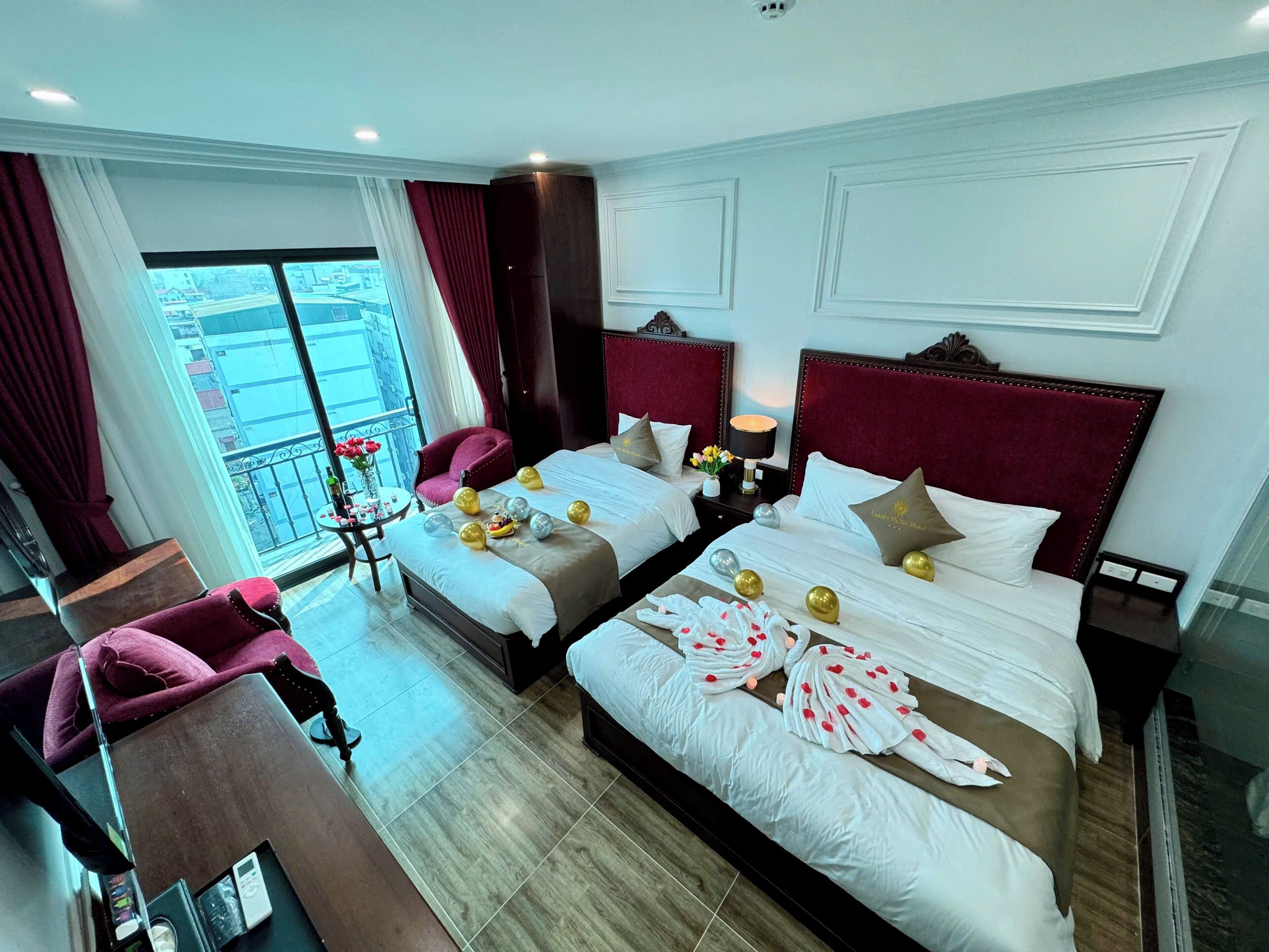 A hotel room with two beds, a sitting area with a chair, a small table with a vase of flowers, and a balcony with a view of buildings.