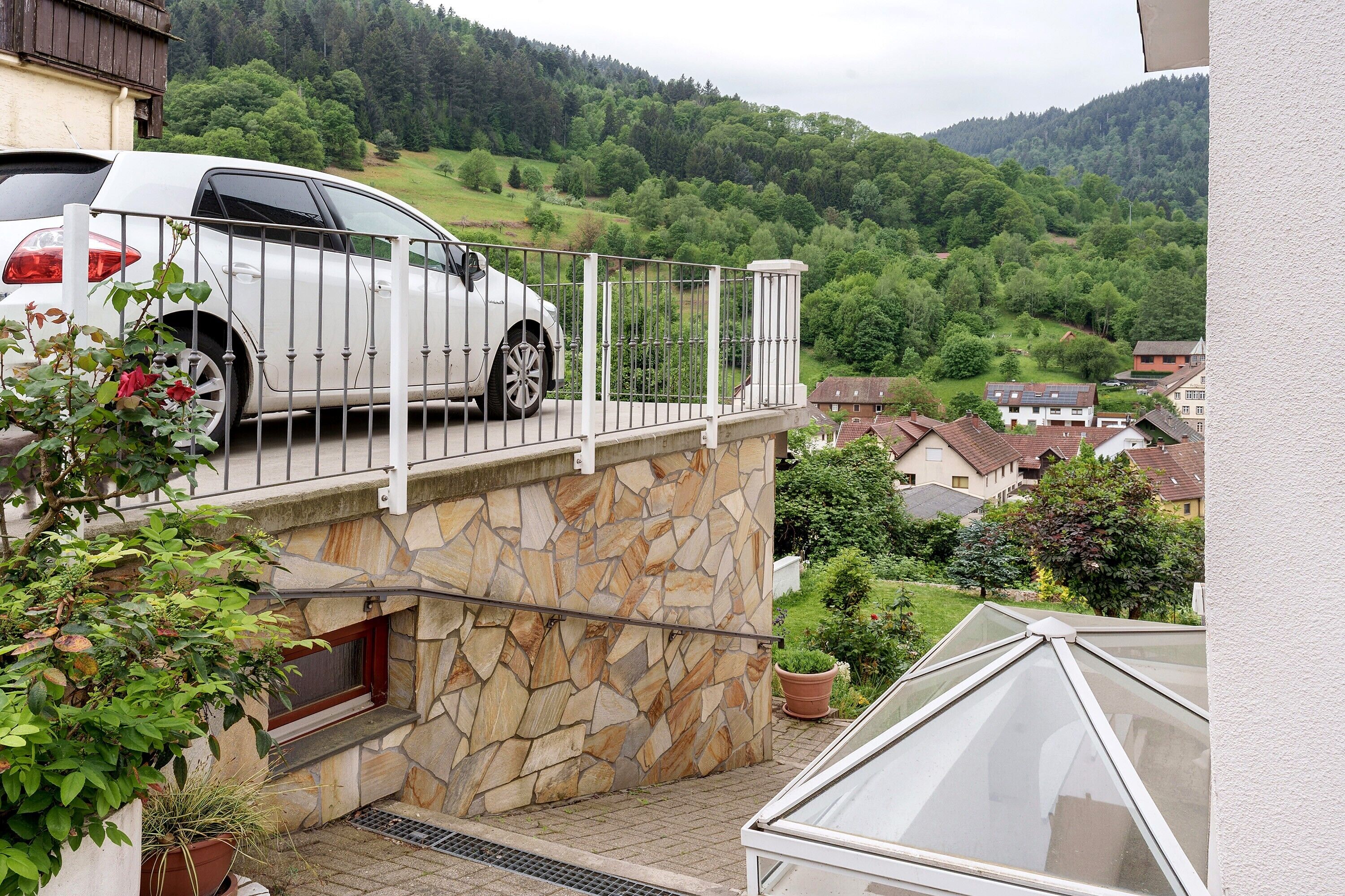 Een witte auto geparkeerd op een balkon, met een stenen muur, een raam en uitzicht op een groene heuvel.