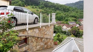 Een witte auto geparkeerd op een balkon, met een stenen muur, een raam en uitzicht op een groene heuvel.