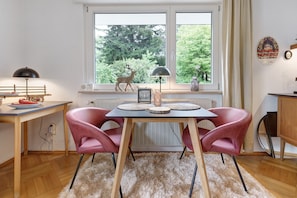 Une pièce avec une table en bois, deux chaises roses, une fenêtre décorée d’un motif de cerf, une lampe et un radiateur.