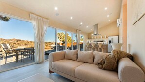 A modern living room with a beige sofa, a coffee table, and a view of a desert landscape through large sliding glass doors. - Wind View Pioneertown (Pioneertown)