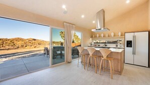 A modern kitchen with a large island, bar stools, a refrigerator, and a view of a desert landscape through the sliding glass door. - Wind View Pioneertown (Pioneertown)