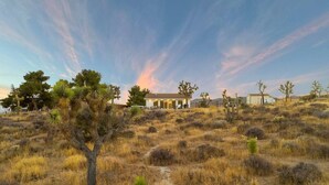 A house with a pathway leading to it, surrounded by dry grassland and sparse trees. - Wind View Pioneertown (Pioneertown)