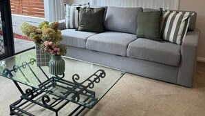 A living room with a grey sofa, a glass coffee table with a vase of flowers, and a view of a garden through a sliding door.