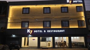 Un bâtiment abritant l’hôtel et le restaurant K7, doté d’une façade moderne, de nombreuses fenêtres et d’une entrée bien visible avec un espace extérieur aménagé avec des sièges.