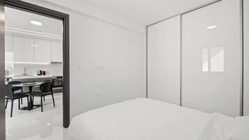 A modern bedroom with a large bed, a sliding closet door, and a view into a kitchen area with a dining table and chairs.