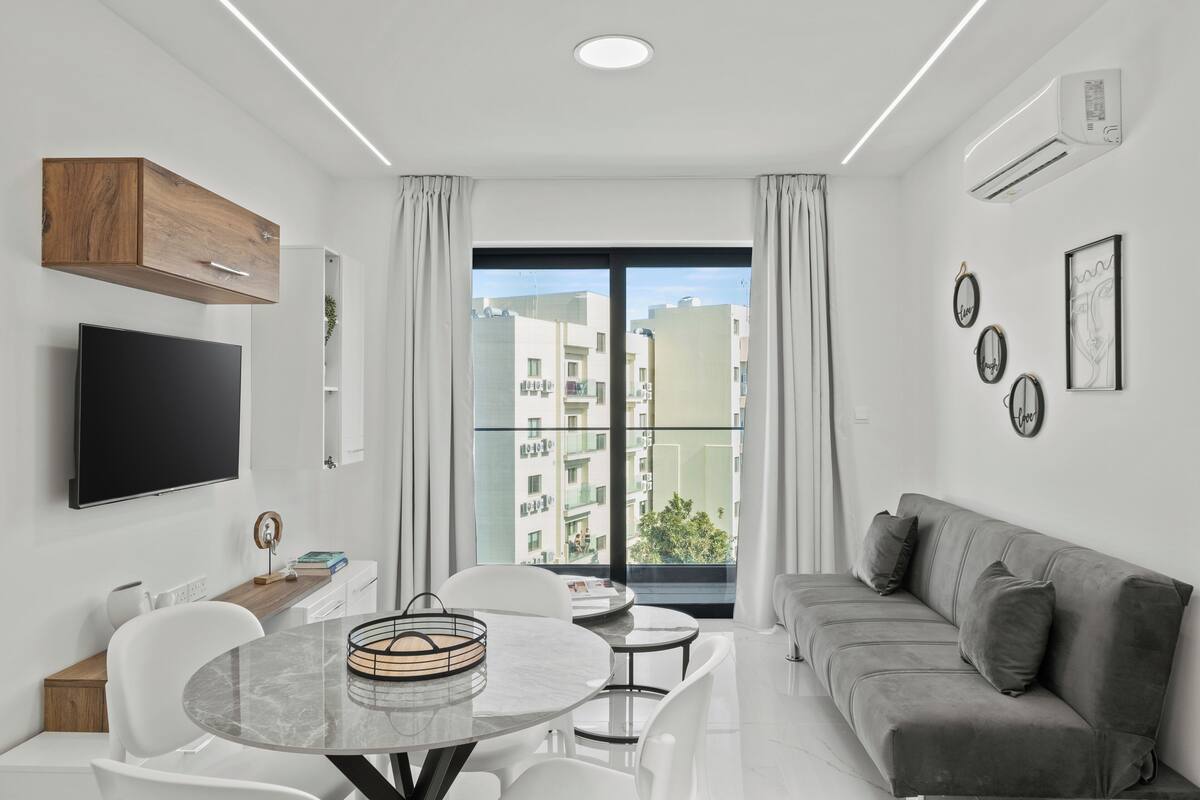 Una sala de estar moderna con un televisor de pantalla plana, un sofá gris, una mesa de comedor con sillas y un ventanal con vistas a la ciudad.
