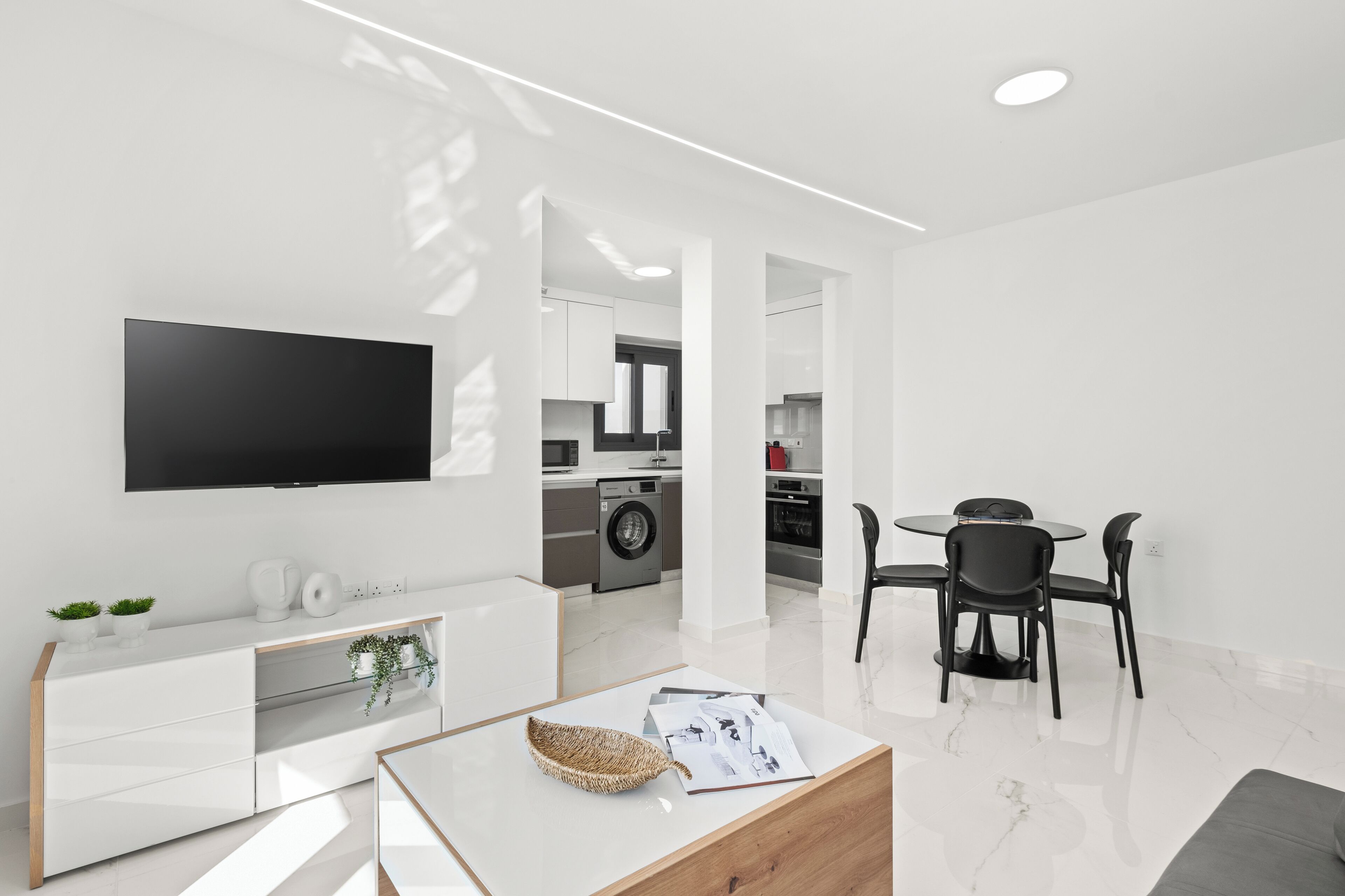 A modern living room with a flat-screen TV, white entertainment unit, and a dining area with a round table and chairs.