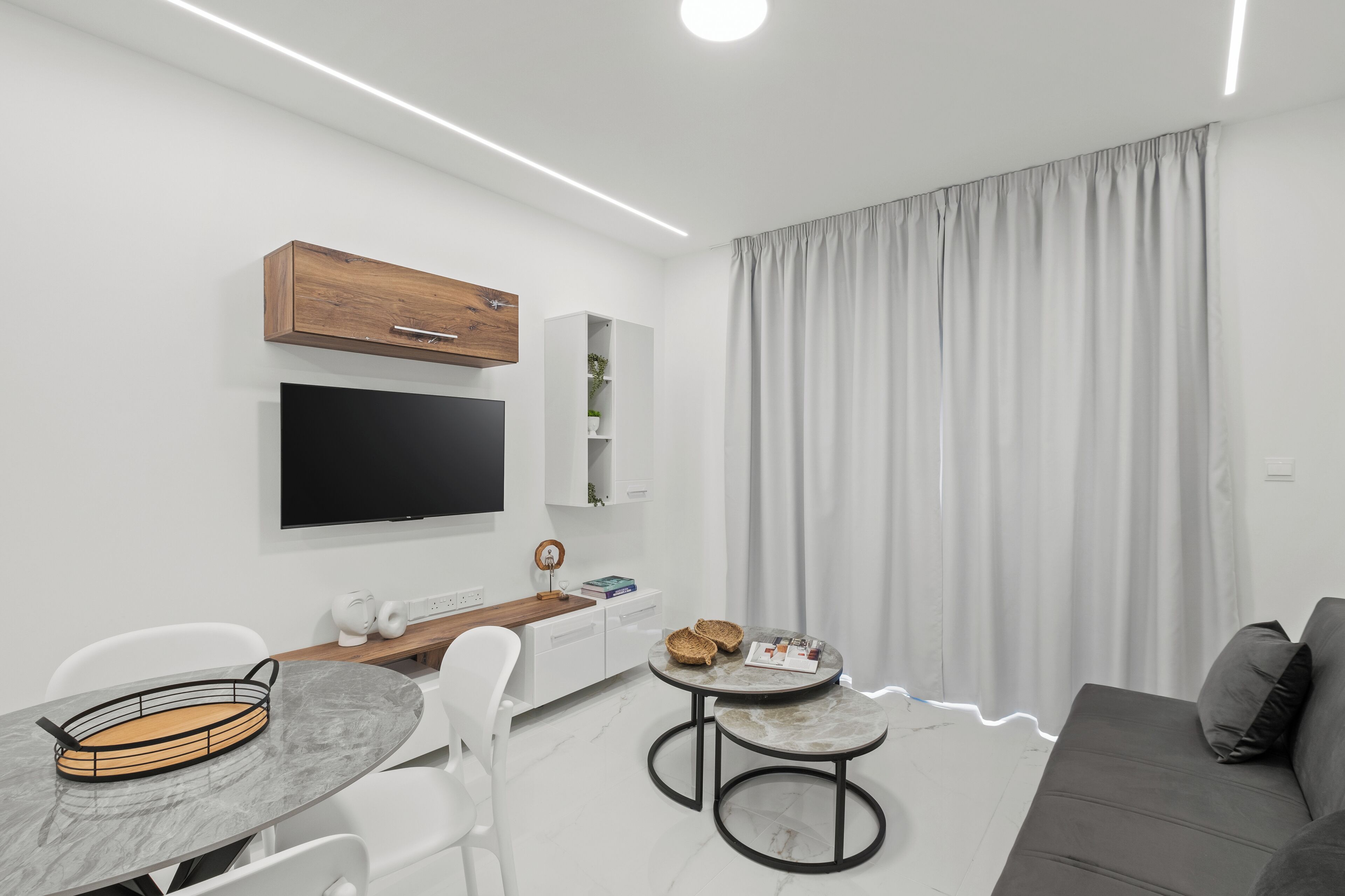 A modern living room with a flat-screen TV, a wooden cabinet, a dining table with chairs, and a gray sofa.