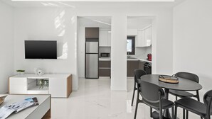 Un espacio habitable moderno y minimalista con una televisión de pantalla plana, una zona de comedor con mesa y sillas redondas, y una cocina con electrodomésticos de acero inoxidable.