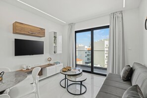 A modern living room with a flat-screen TV, a dining table, a sofa, and a large window offering a city view. - Maestro City Apartments (Larnaca)