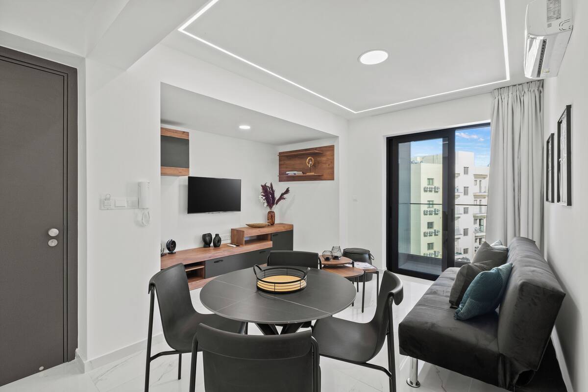 Una sala de estar moderna con una mesa de comedor redonda, sillas negras, un sofá gris y un gran ventanal con vista a la ciudad.