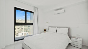 Un dormitorio moderno con un ventanal grande, una cama, una mesita de noche con lámpara y un aire acondicionado de pared.