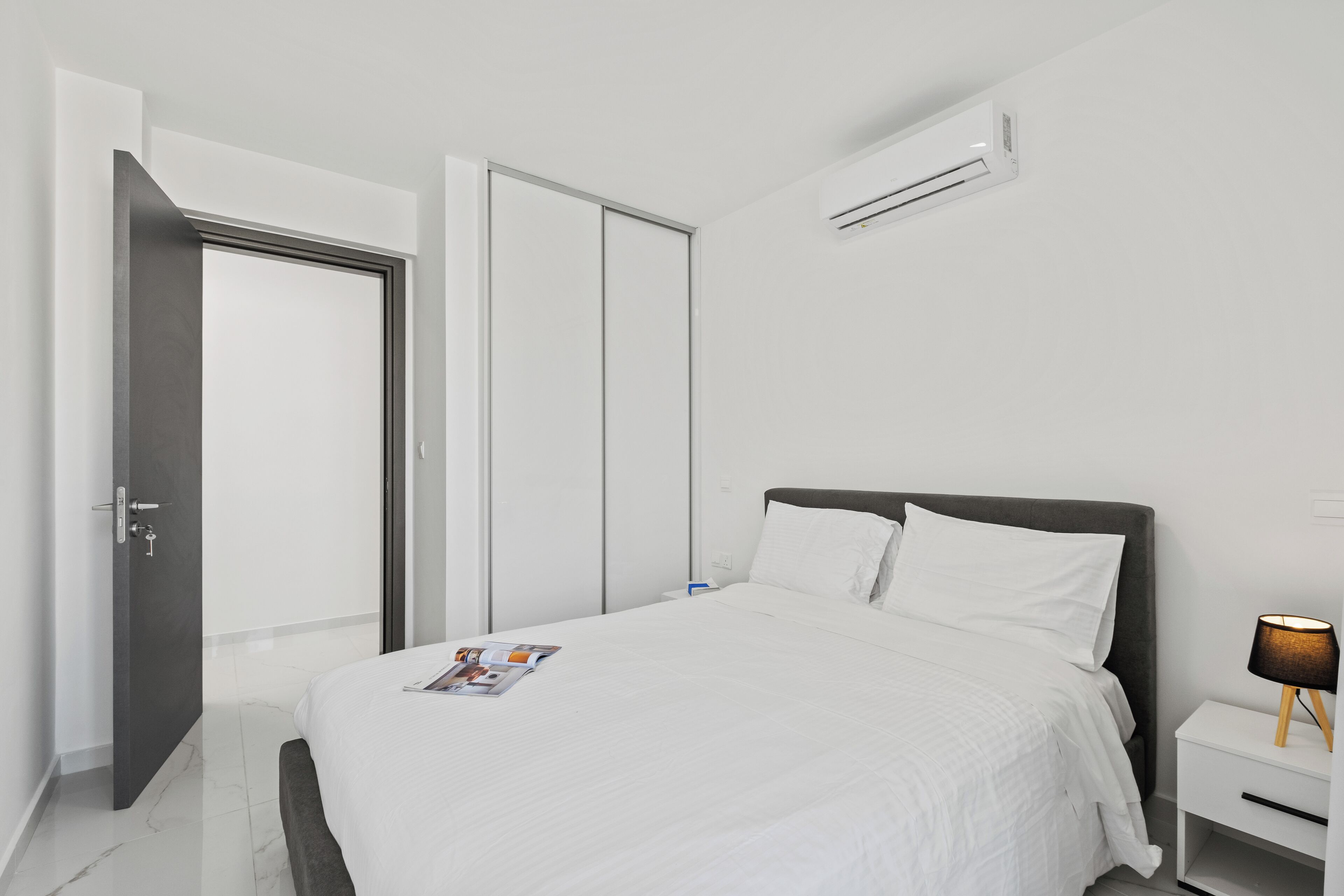 A modern hotel room with a bed, a nightstand, a lamp, and an air conditioning unit.