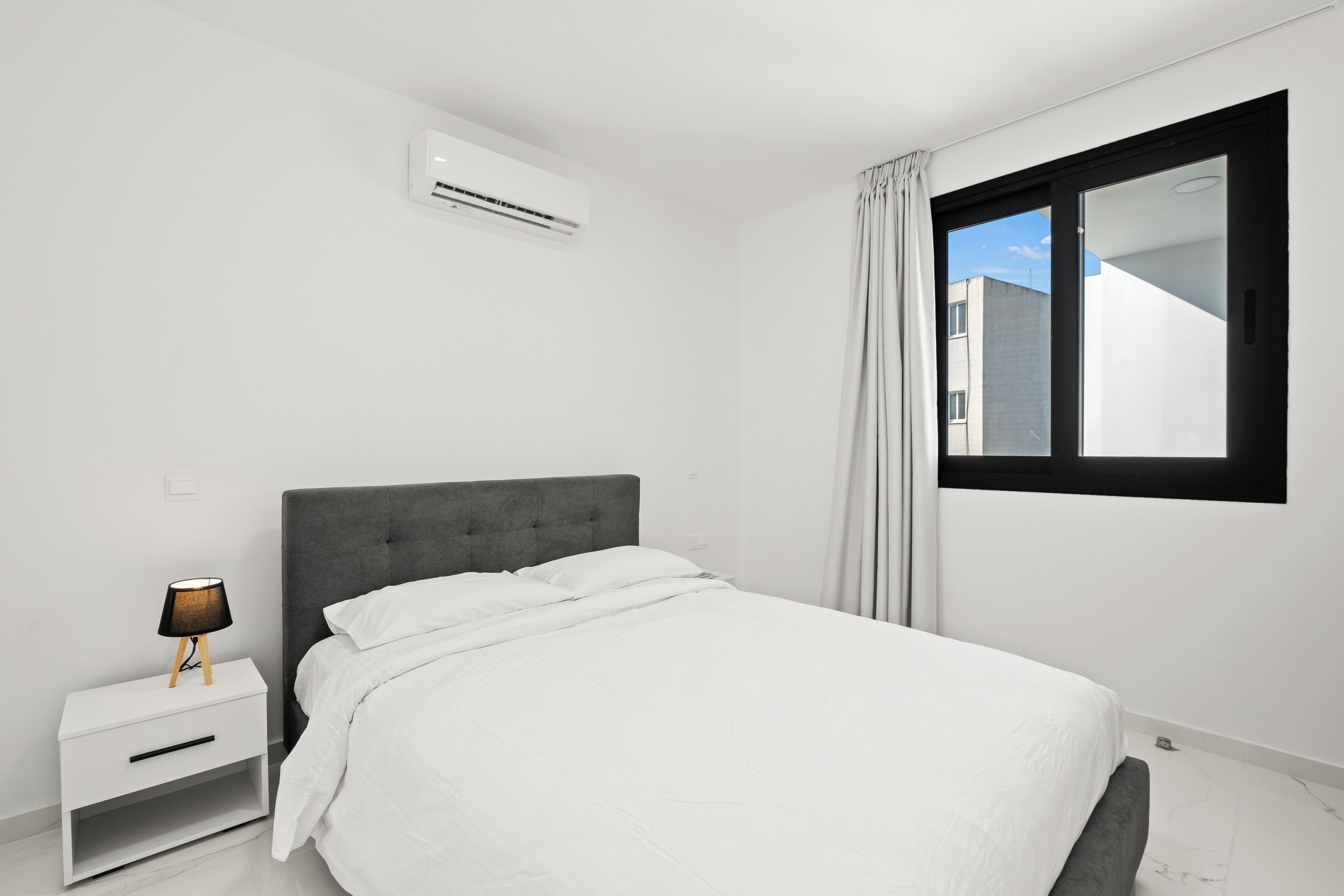 A modern bedroom with a grey upholstered headboard, white bedding, a bedside lamp, and a window with curtains.