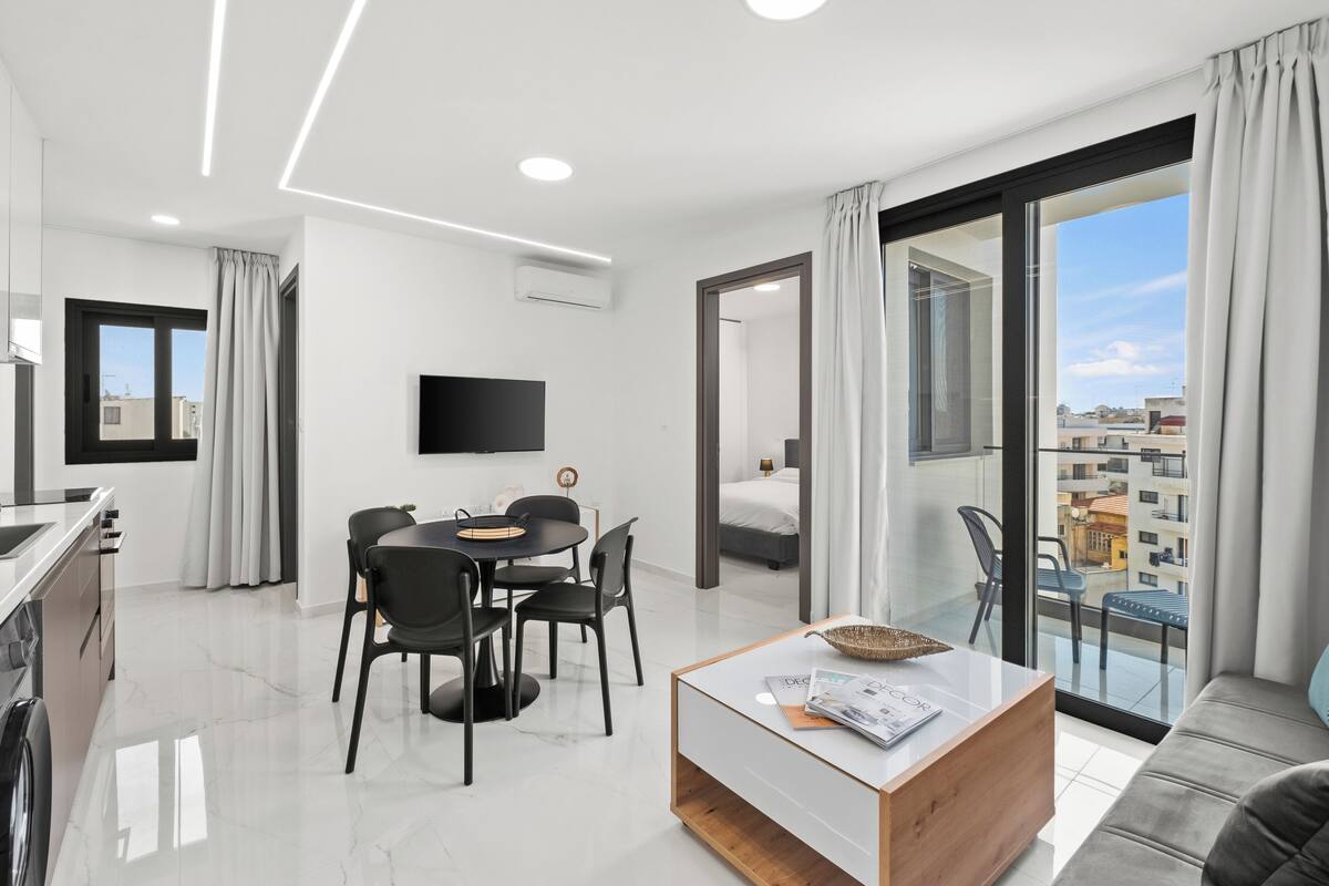 Una habitación de hotel moderna con comedor, sofá, televisor de pantalla plana y balcón con vistas a la ciudad.