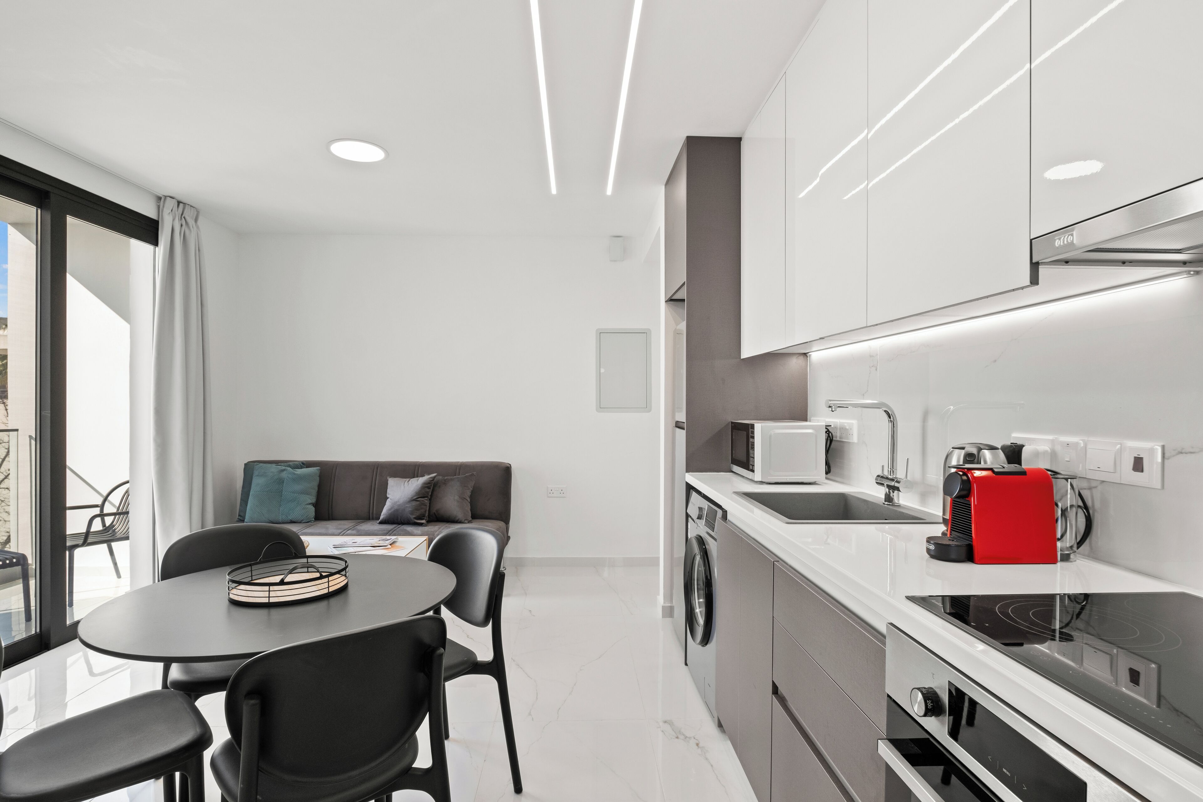 A modern kitchen with a microwave, coffee machine, and a washing machine. There is a dining area with a table and chairs.