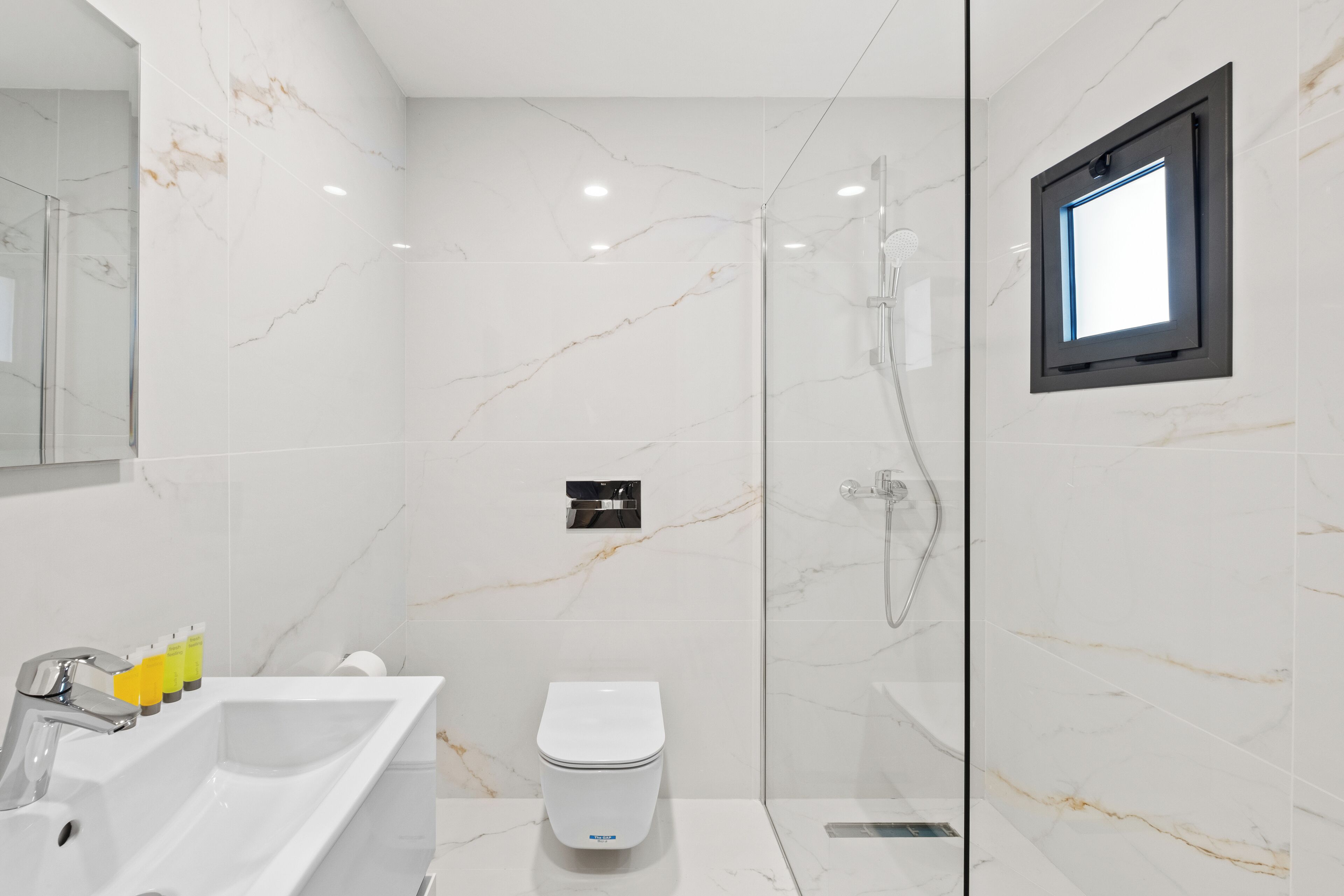 A modern bathroom with a white marble wall, a glass shower enclosure, a white toilet, and a built-in window.