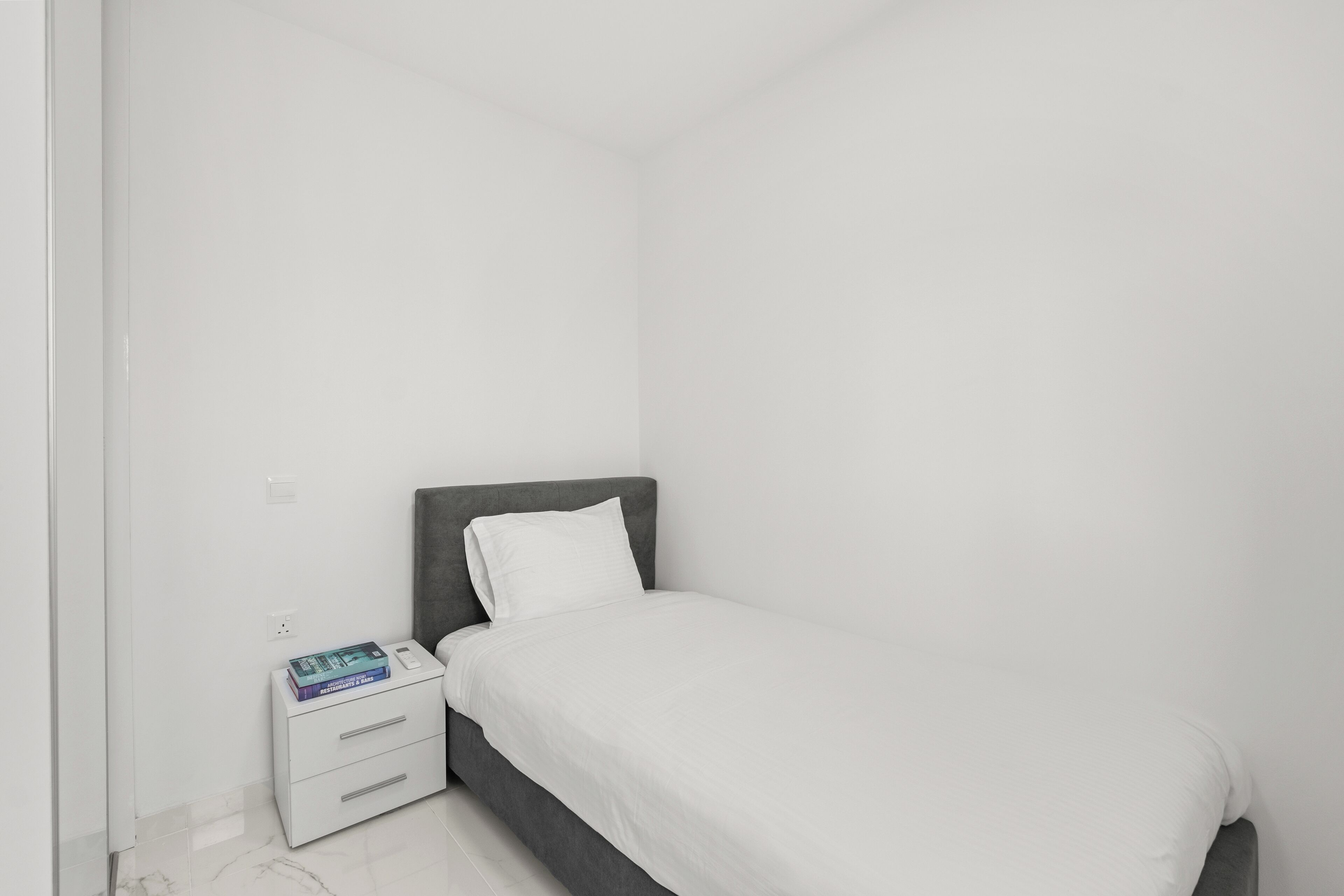 Un dormitorio minimalista con una cama individual, una mesita de noche y un pequeño conjunto de cajones.