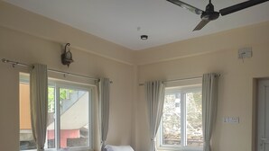 Una camera d'albergo con tre letti, una ventola a soffitto e ampie finestre con tende.