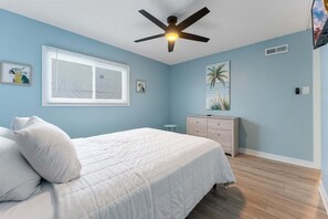 Une chambre à coucher comprenant un lit, un ventilateur de plafond, une fenêtre avec des stores, une table de chevet, une commode et des œuvres d’art murales.