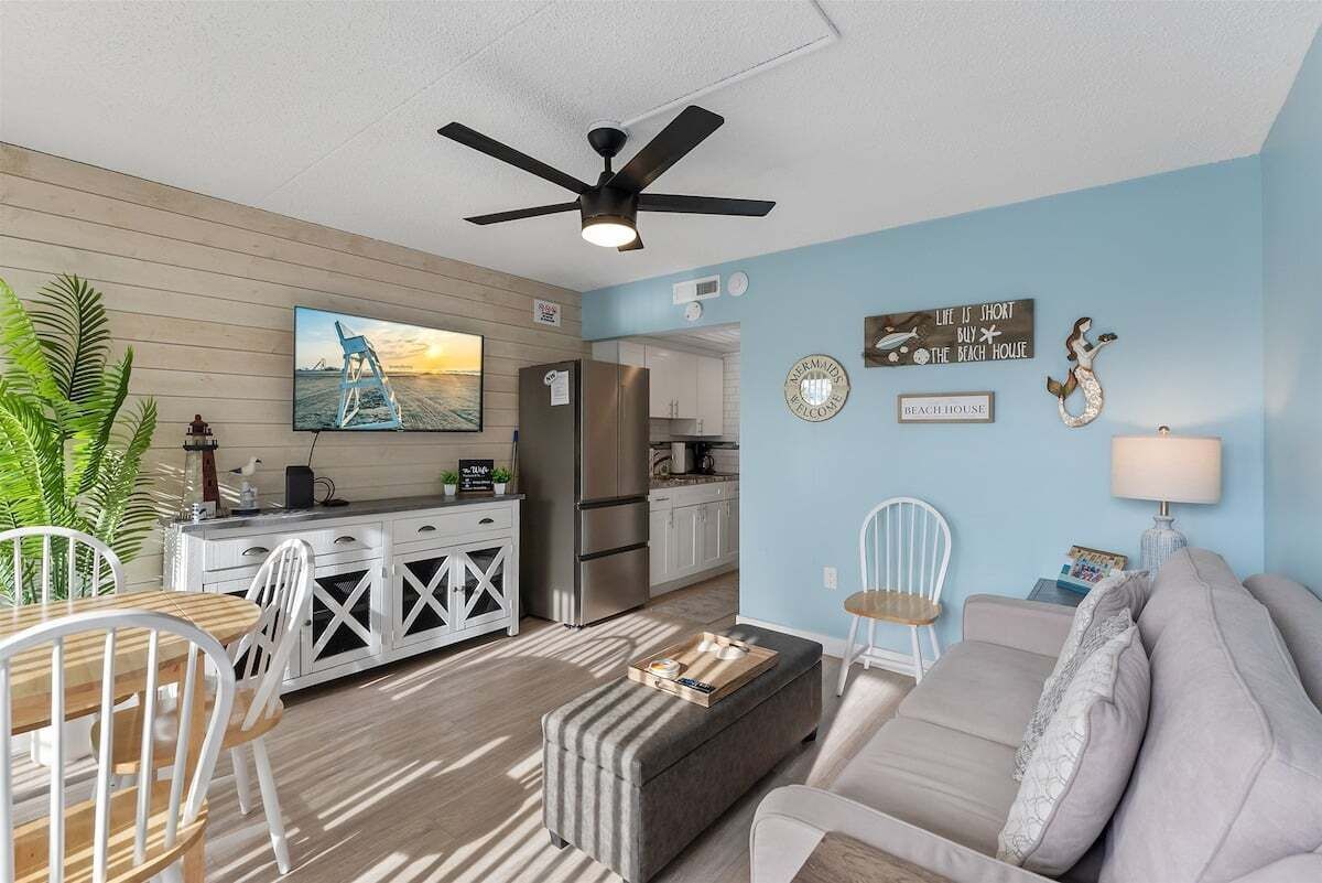 A modern living room with a sofa, a dining table with chairs, a kitchen area, and a ceiling fan.