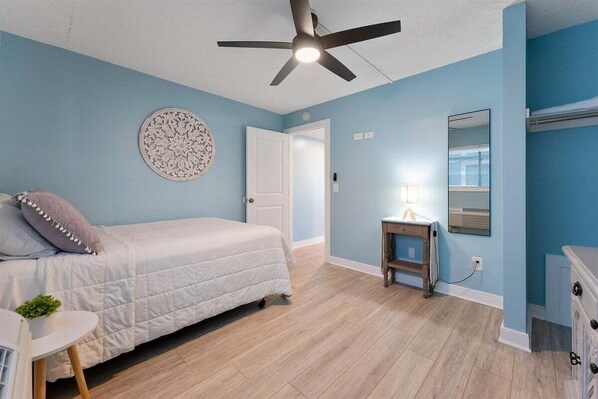 A bedroom with a bed, a ceiling fan, a bedside table with a lamp, a wall decoration, and a dresser.