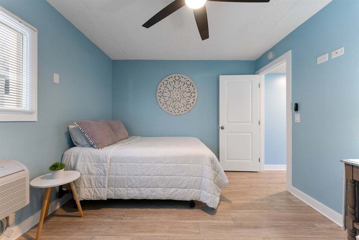 Ein Schlafzimmer mit einem Bett, einem Deckenventilator, einer an der Wand befestigten Dekorplatte, einer Tür und einem kleinen Tisch mit einer Pflanze.