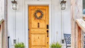 Una porta de fusta amb una corona decorativa, emmarcada per dues plantes en testos i una cadira.