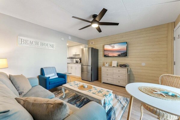 Ein Wohnzimmer mit einer Couch, einem blauen Sessel, einem Couchtisch, einem Deckenventilator, einer Küchenzeile mit Kühlschrank und einem Fernseher an der Wand.