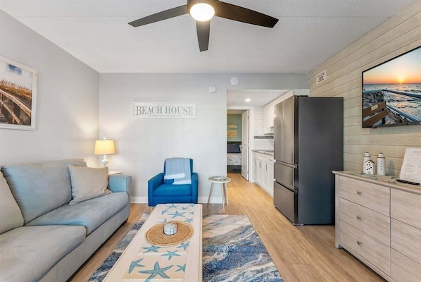 A living room with a sofa, a blue chair, a coffee table with star decorations, a ceiling fan, and a kitchen area with a refrigerator and cabinets.