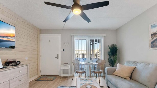 A modern living room with a ceiling fan, a sofa, a dining table with chairs, a TV mounted on the wall, and a window with blinds.