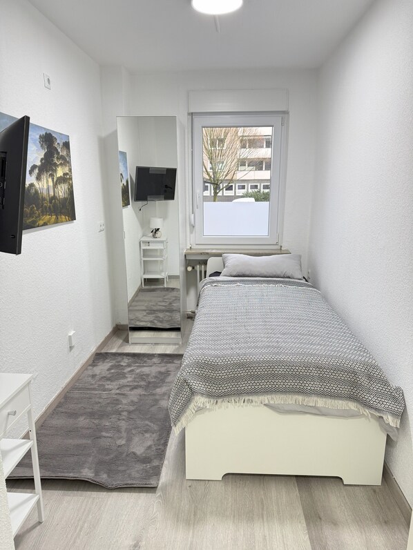 A small, well-lit bedroom with a bed, a desk, a chair, a TV, and a window with a view of buildings. - #3 Top Room: centrally located & comfortable, shared bathroom (Herne)