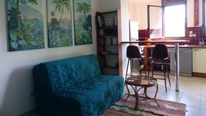 A living room with a blue sofa, two chairs, a small table, and a colorful rug. There are three large tropical-themed paintings on the wall. - Attractive T2 apartment on the Saint Pierre waterfront in an ideally located building. (Saint-Pierre)