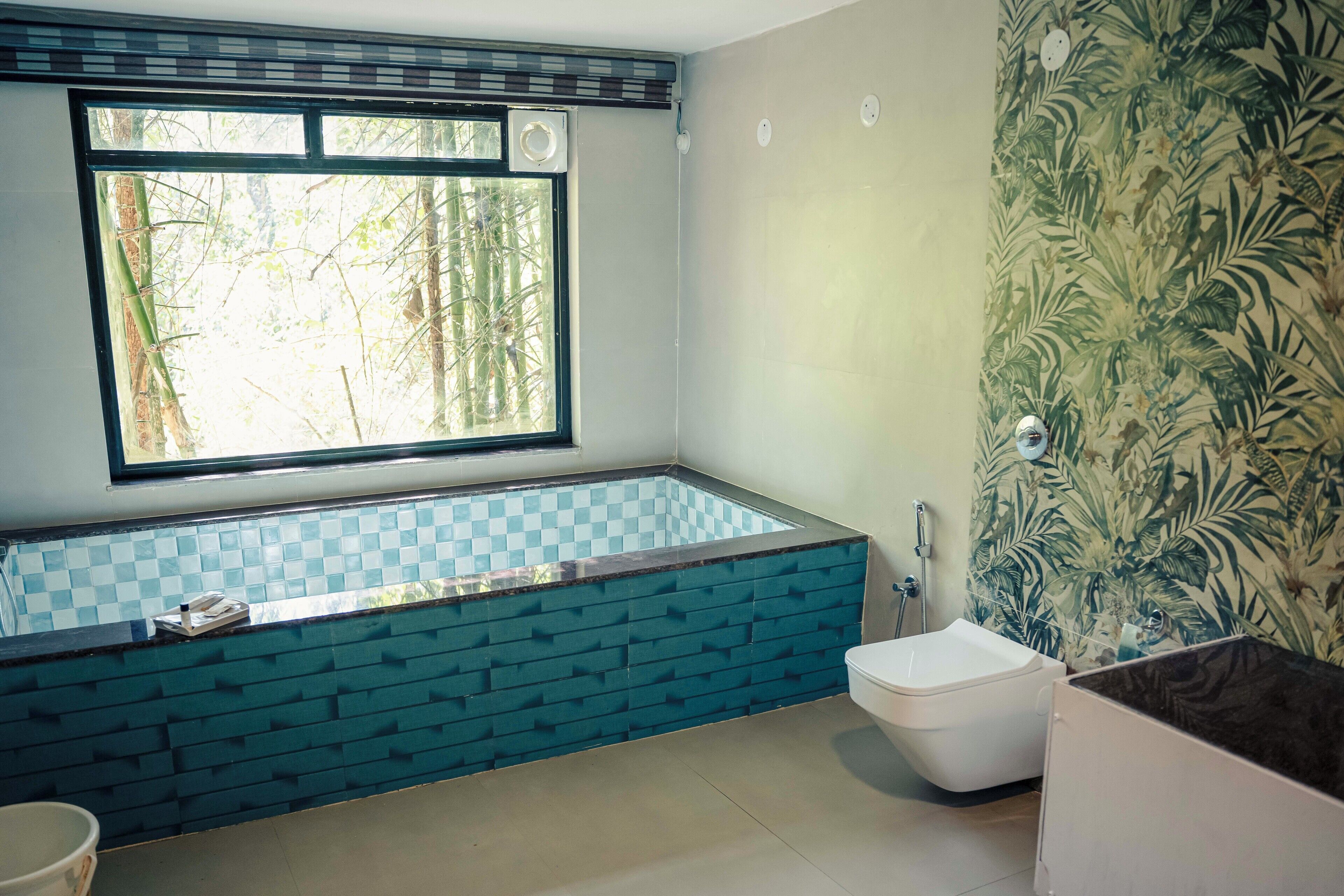 A bathroom with a checkered tile bathtub, a toilet, and a window with a view of trees.