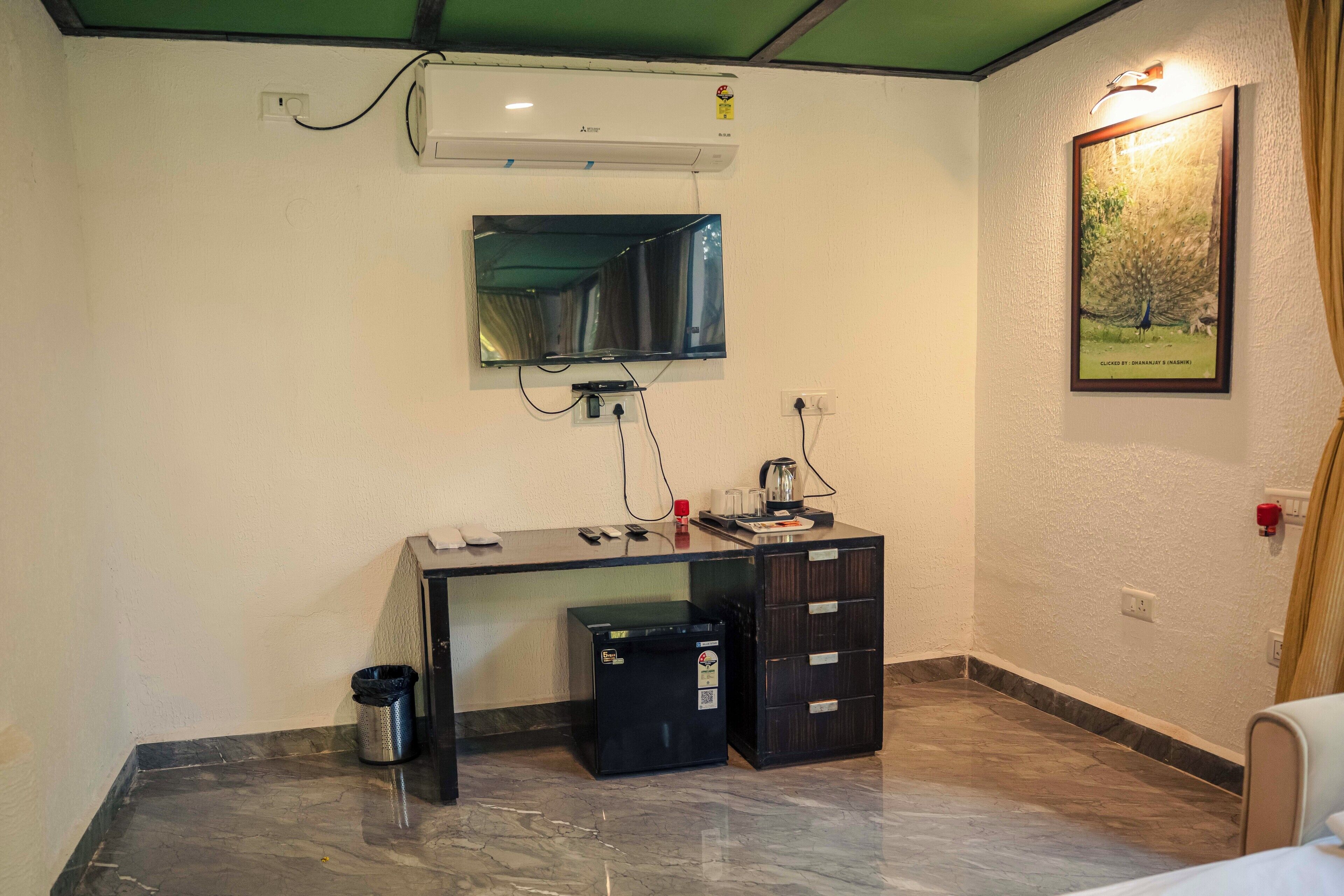 Habitación de hotel compacta con escritorio, refrigerador pequeño, cesto de basura, televisor montado en la pared y un cuadro enmarcado.