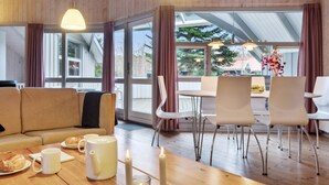 A modern living room with a wooden floor, a sofa, a dining table with chairs, and large windows with curtains.