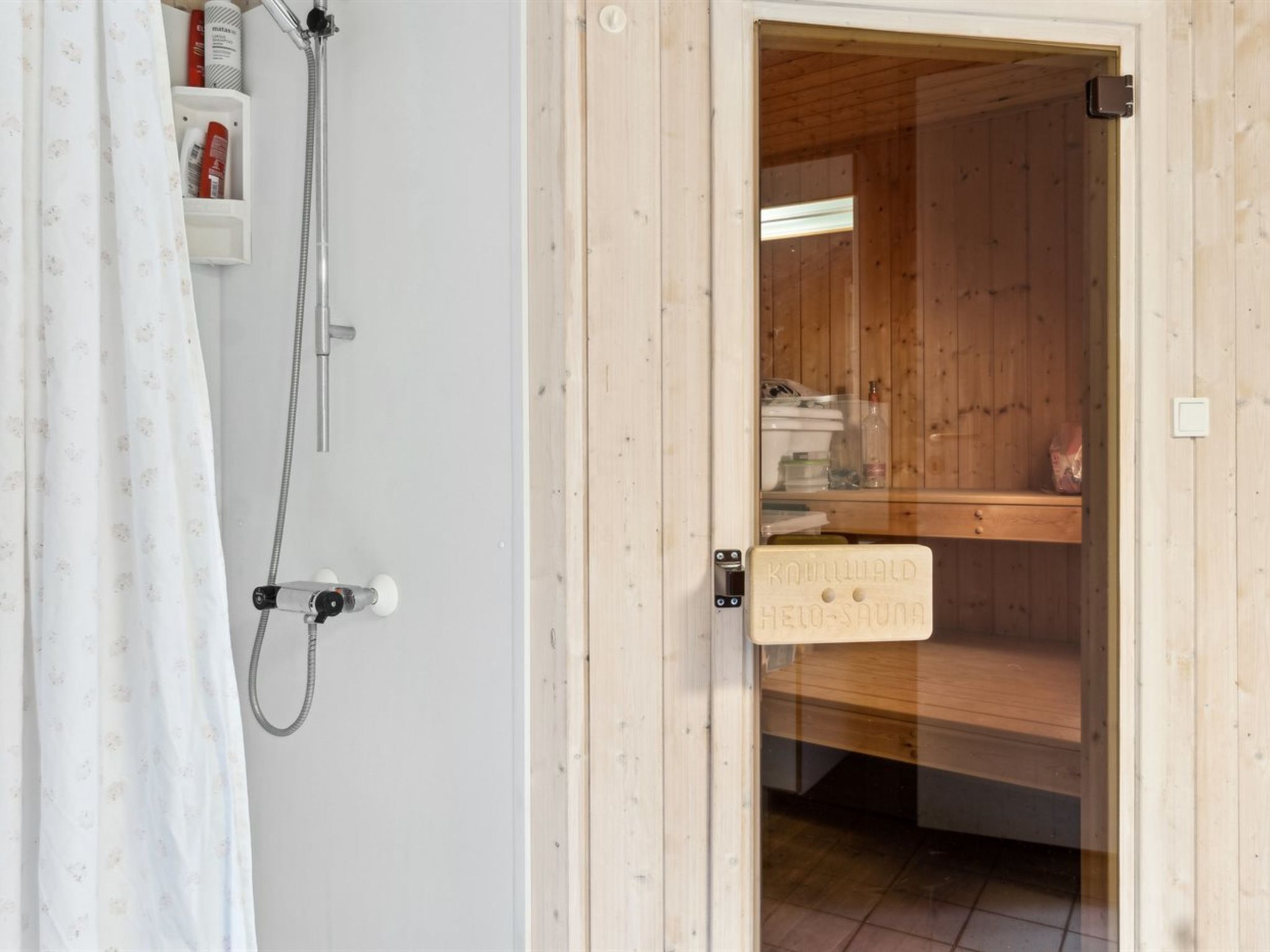 Un sauna de madera con puerta de madera, zona de ducha con cabezal y estante con artículos de aseo.