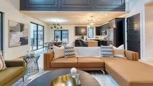 A modern living room with a brown leather sofa, a round coffee table, and a dining area in the background. - Indoor Pool, Hot Tub, Firepit, Theater | All-Season Retreat | Kings Crown (Gatlinburg)