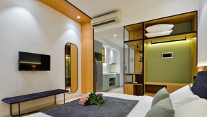 Una habitación de hotel moderna con cama, un banco, televisor, kitchenette y un mueble de almacenamiento con almohadas.