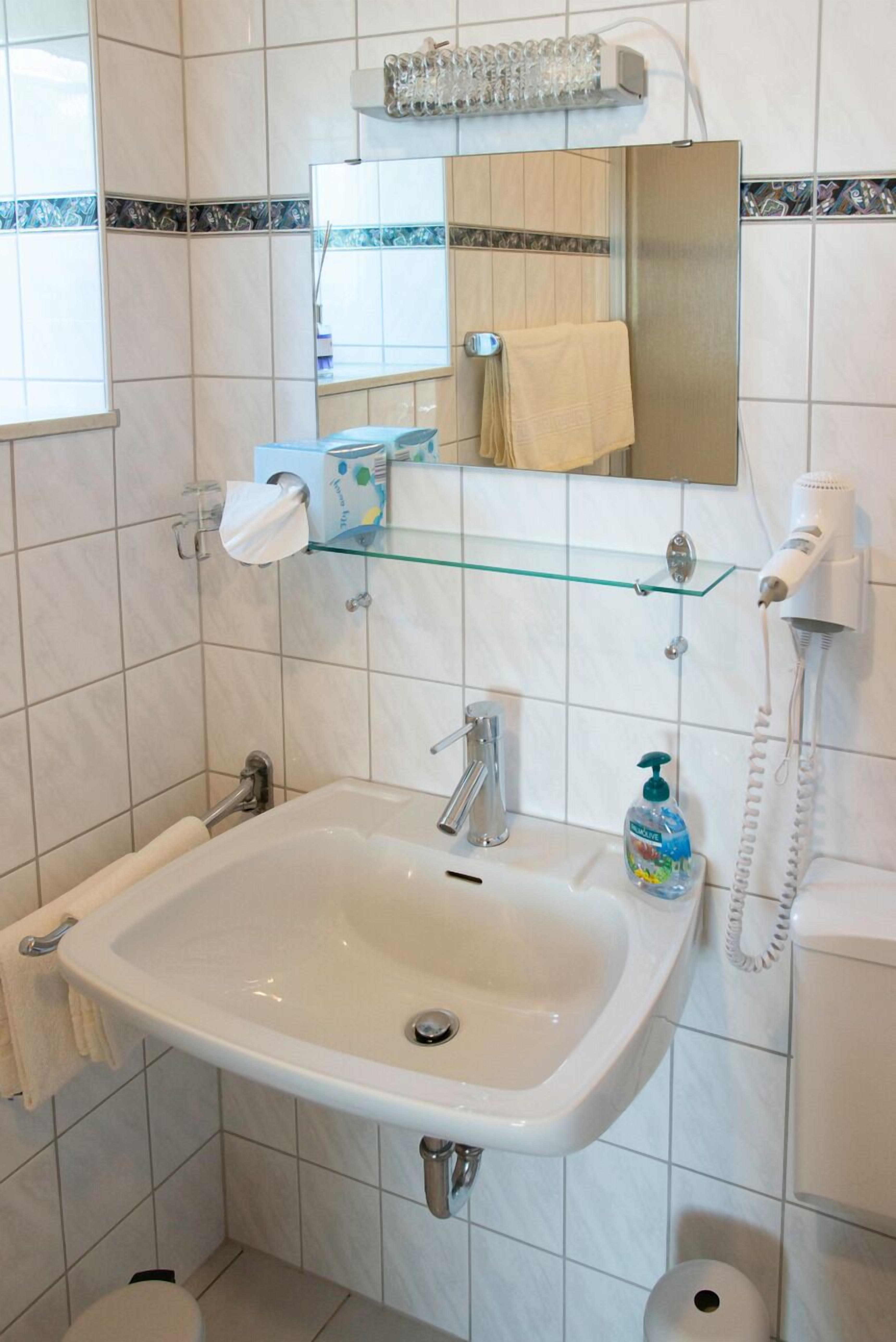 Un baño con lavamanos blanco, un espejo, repisas de vidrio con artículos de aseo y un secador de pelo.