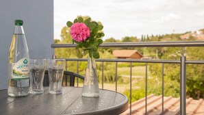 A round table with a bottle of Hochwa water, two glasses, and a vase with a pink flower on a balcony with a metal railing and a view of greenery and buildings. - Birkenhof Family Lodge  (Rieschweiler-Mühlbach)